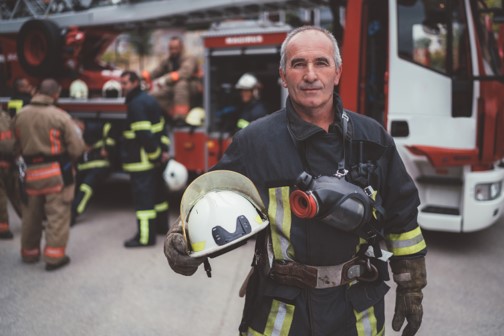 Retired firefighter in uniform holding his helmet, standing confidently in front of a fire truck with fellow firefighters in the background. Represents Peak 65 retirees seeking reliable retirement income solutions. Ideal visual for annuity planning, lifetime income, and financial security in retirement.